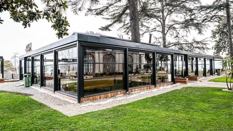 Bahçe içinde giyotin cam ile kapatılmış restoran; siyah alüminyum profiller ve tam boy cam paneller.