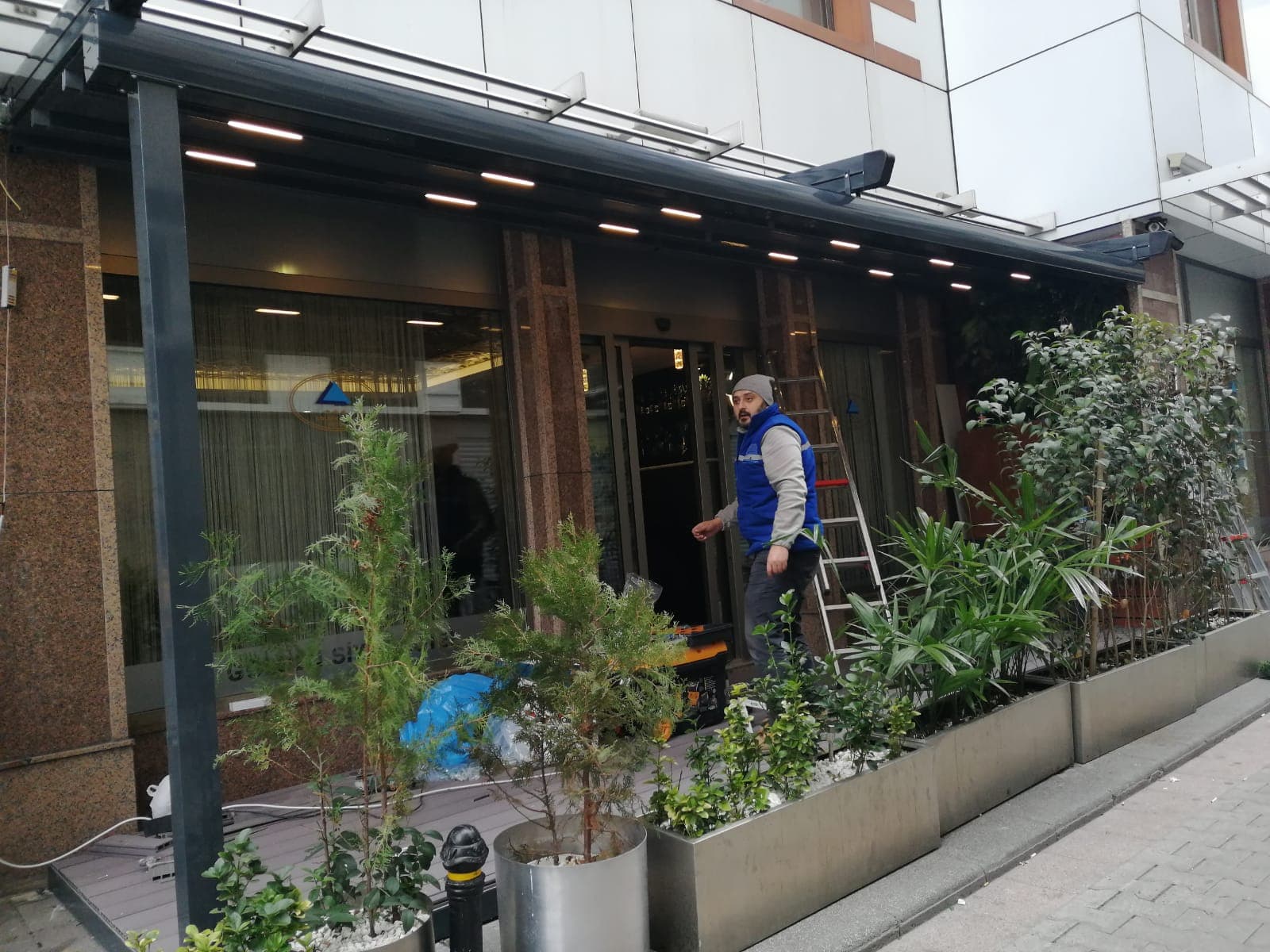Ticari bir cephede devam eden montaj çalışması; üstte LED aydınlatmalı alüminyum konstrüksiyon ve montaj ekibinden bir çalışan.